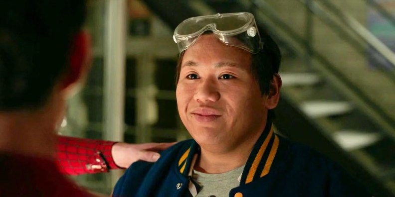 A young man wearing a blue varsity jacket and safety goggles, smiling and engaging with another person.
