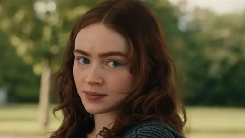 A close-up of a young woman with wavy, reddish-brown hair, looking over her shoulder with a thoughtful expression, set against a natural background of greenery.