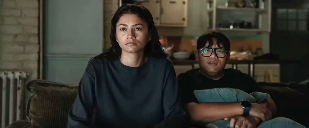 A woman with long hair and a serious expression sitting next to a young man wearing glasses, both watching something on a screen. The setting is a cozy living room with a couch and kitchen in the background.