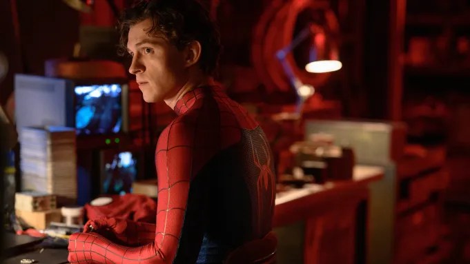 A young man in a Spider-Man costume sitting at a desk with monitors and equipment in a dimly lit room.