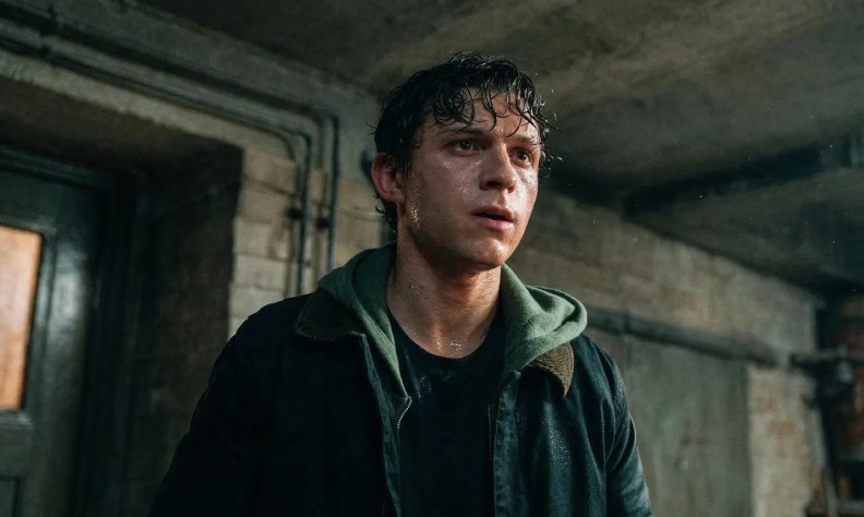 A young man with wet hair and a serious expression stands in a dimly lit room, wearing a black jacket over a green hoodie.