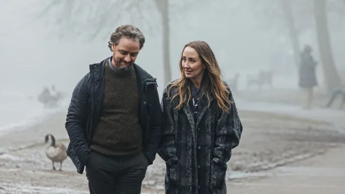 A man and a woman stroll along a foggy pathway by the water, engaging in conversation. A duck is seen nearby, and the background features blurred figures and trees.