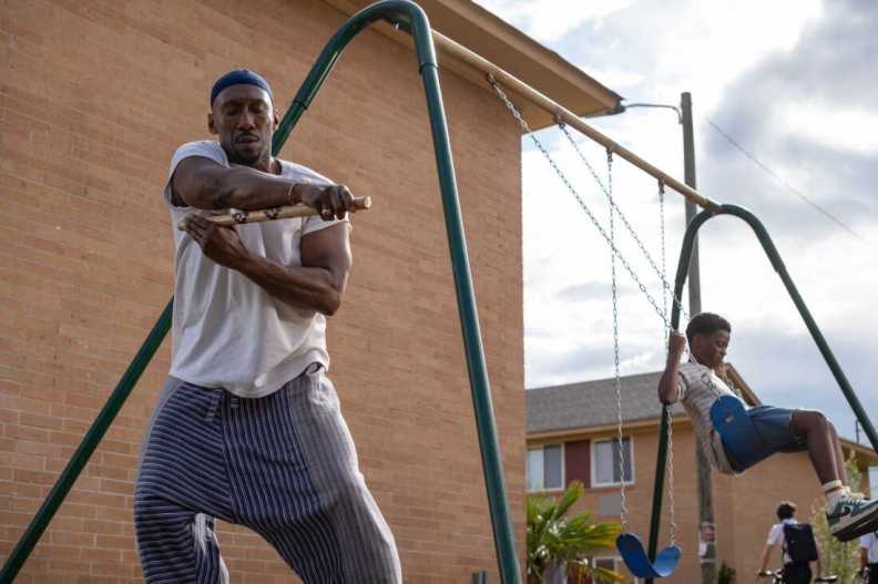 A man in a white shirt and striped pants is demonstrating a movement with a stick while standing next to a swing set, where a boy in a patterned shirt is happily swinging. The background features a brick building and a clear sky.