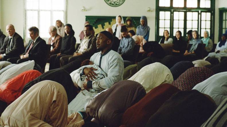 A diverse group of individuals gathered indoors, participating in a prayer service. Some people are seated while others are bowing in reverence. The atmosphere is serene and reflective.